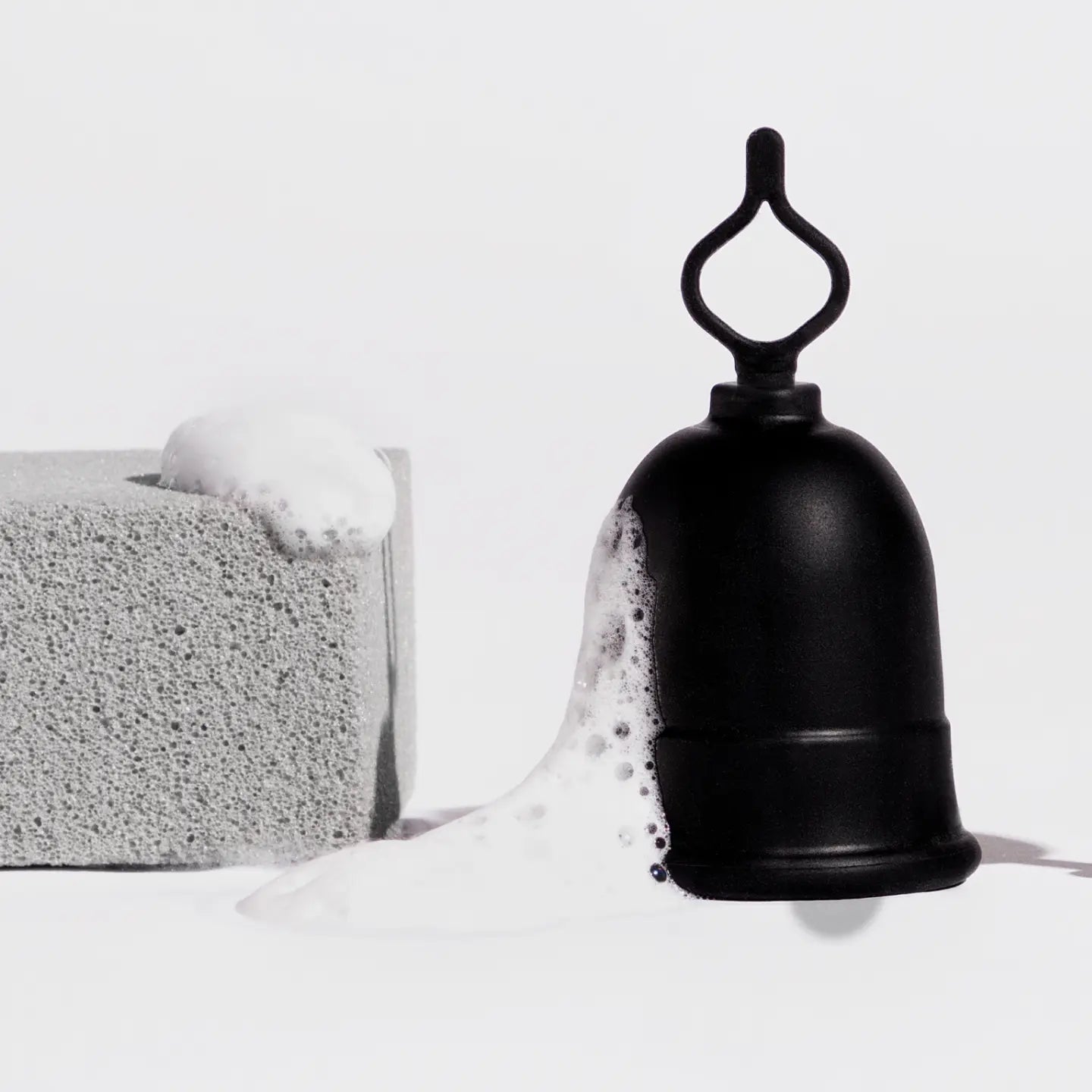 Menstrual cup with loop stem shown next to pumice block and soap suds